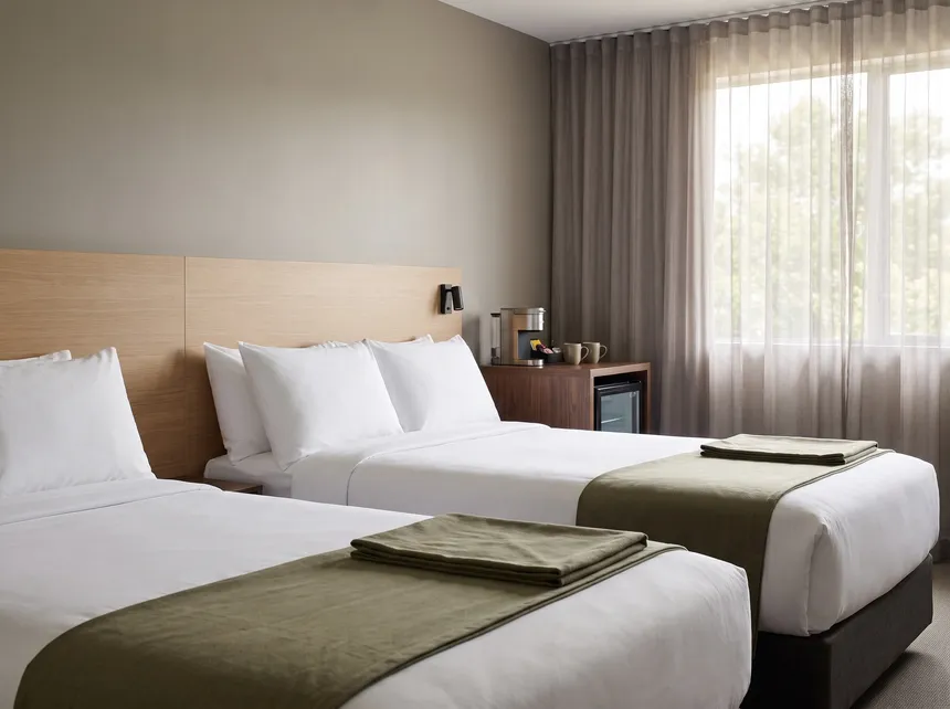 Illustration: a hotel room with two made queen beds, olive throws, a desk with a coffee maker and a mini fridge.